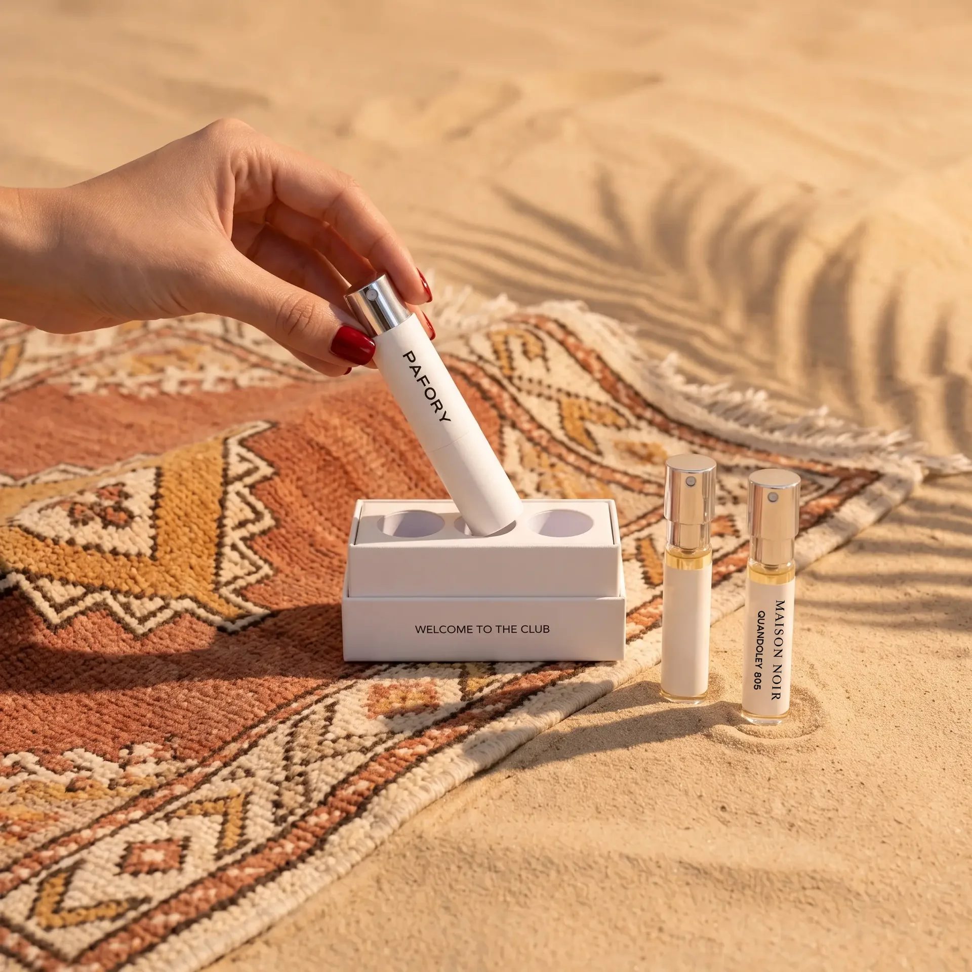 Hand placing the white PAFORY case into the Member Box on a Berber rug in the desert sand, next to the 8 ml Maison Noir Quandoley 805 scent